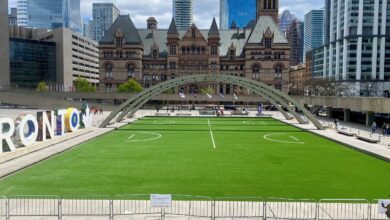 Toronto sign and artificial turf field at Nathan Phillips Square with Old City Hall and tall glass skyscrapers in the background.