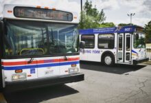 Front-left view of a white transit bus with red/blue stripes and the sign '15 via CITY CENTRE', parked beside a York Region Transit bus labeled 'MOVING YORK' on a sunny day.