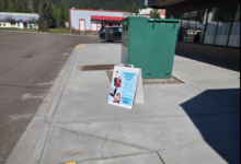 A-frame sidewalk sign advertising a community event beside a green utility cabinet in front of a storefront.3ea