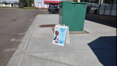 A-frame sidewalk sign advertising a community event beside a green utility cabinet in front of a storefront.3ea