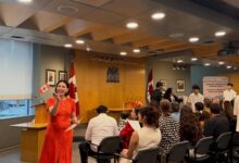 Woman in an orange dress speaks to an audience at a formal ceremony, with Canadian flags and a wooden podium nearby.