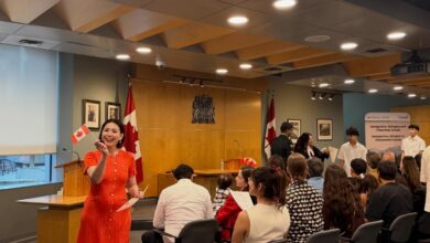 Woman in an orange dress speaks to an audience at a formal ceremony, with Canadian flags and a wooden podium nearby.