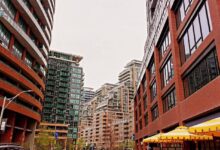 City street lined with tall modern buildings, pedestrians walking along a sidewalk and a cafe with yellow umbrellas.