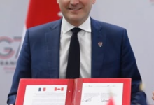 Man in a blue suit and tie sits at a desk, signing a document in a red binder, with French and Canadian flags visible on the pages and a Canadian flag backdrop.