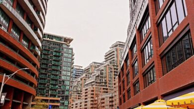 City street lined with tall modern buildings, pedestrians walking along a sidewalk and a cafe with yellow umbrellas.