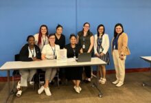 Nine people posing for a group photo around a conference table in a room with a blue wall; two laptops and a flyer visible on the table, everyone wearing lanyards.