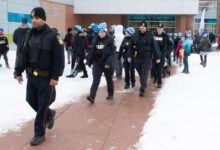 Line of police officers in winter uniforms walking on a snowy sidewalk outside a brick building with a crowd in the background.