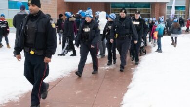 Line of police officers in winter uniforms walking on a snowy sidewalk outside a brick building with a crowd in the background.