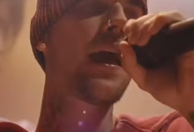 Male singer wearing a red beanie and hoodie, singing into a microphone on stage