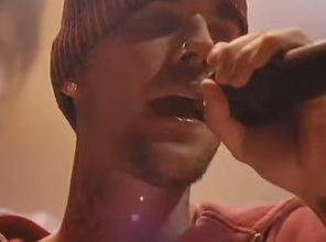 Male singer wearing a red beanie and hoodie, singing into a microphone on stage