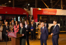 Dignitaries and staff gather at a podium for an unveiling beside red 100% electric buses in a depot setting.