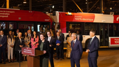 Dignitaries and staff gather at a podium for an unveiling beside red 100% electric buses in a depot setting.