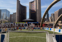 Portable soccer goal on a green turf field at a street tournament, with tall city skyscrapers and a curved arch in the background.