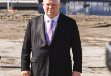 Older man in a black suit with a purple tie stands on a sandy waterfront area, with a marina and boats in the background.