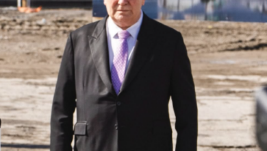 Older man in a black suit with a purple tie stands on a sandy waterfront area, with a marina and boats in the background.