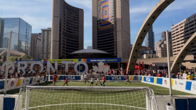 Portable soccer goal on a green turf field at a street tournament, with tall city skyscrapers and a curved arch in the background.