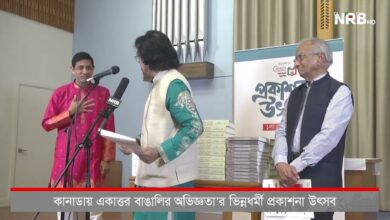 Man in a pink kurta speaks at a microphone on stage, while a man in teal with a beige vest holds papers, with stacks of books and a banner in the background.