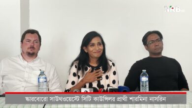 Three panelists at a press conference sit behind a table with microphones; the woman in the middle speaks while the two men listen.