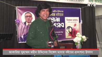Man in a green kurta speaking into a microphone at a stage, with a purple backdrop banner and a book cover visible behind him.