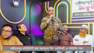 A man stands with a microphone addressing an audience during a panel discussion, with four men seated on a colorful stage behind him and a Bengali banner visible to the right.