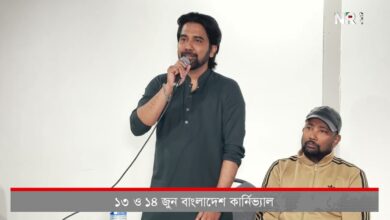 Man standing and speaking into a microphone at a panel, with a seated man in a cap beside him; Bengali news ticker runs across the bottom.