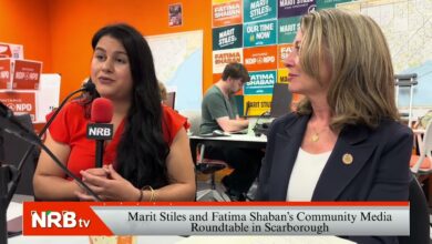 Two women in a bright orange studio; a reporter with a red NRB mic speaks while another woman in a blazer listens, campaign posters in the background.