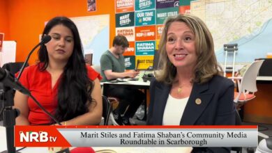 Two women sit at a talk-show table in a bright orange studio; one in red sits with a microphone, the other in a blazer smiles as posters and a map fill the background.