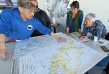 Group of adults gathered around a large map on a table; one man in a blue hoodie points at a route as others study.