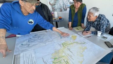 Group of adults gathered around a large map on a table; one man in a blue hoodie points at a route as others study.