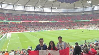 Three fans stand in a packed stadium holding a Canadian flag, with a large video screen overhead and a green soccer pitch behind them.
