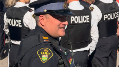 Police officer in uniform smiling among a crowd of officers wearing 'POLICE' vests at an outdoor event, with an ambulance and trees in the background.