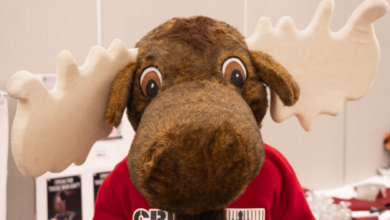 Plush moose mascot wearing a red Crime Stoppers shirt indoors at a booth or event.
