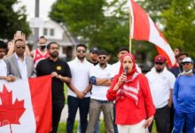 Woman in a red hijab speaks into a microphone at a daytime outdoor rally, with a Canadian flag and a crowd behind her.