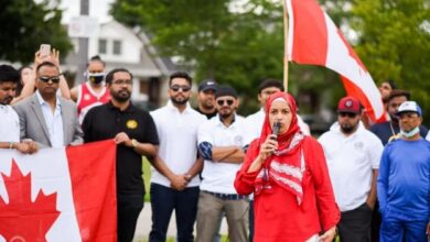 Woman in a red hijab speaks into a microphone at a daytime outdoor rally, with a Canadian flag and a crowd behind her.