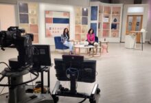 TV studio set with two women hosts seated on blue and pink chairs, beauty bar backdrop; foreground shows cameras and monitors.