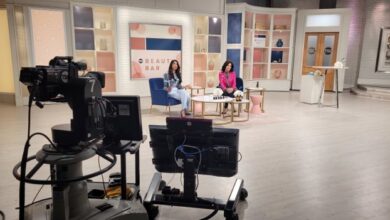 TV studio set with two women hosts seated on blue and pink chairs, beauty bar backdrop; foreground shows cameras and monitors.