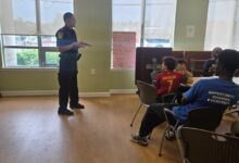 Police officer standing and talking to a small group of students seated around tables in a bright room with large windows, giving a presentation or talk.