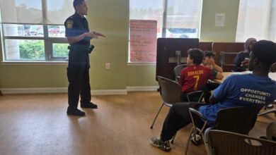 Police officer standing and talking to a small group of students seated around tables in a bright room with large windows, giving a presentation or talk.