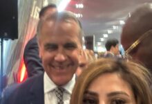 Smiling man in a navy suit and patterned tie posing for a selfie with a woman in the foreground at a crowded indoor event hallway.