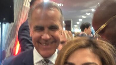 Smiling man in a navy suit and patterned tie posing for a selfie with a woman in the foreground at a crowded indoor event hallway.
