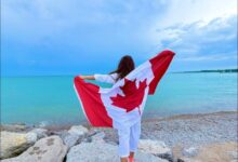 Person standing on rocky shoreline at the beach, draped in a Canadian flag that stretches outward in the breeze toward the turquoise water.