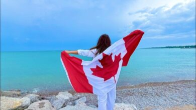 Person standing on rocky shoreline at the beach, draped in a Canadian flag that stretches outward in the breeze toward the turquoise water.